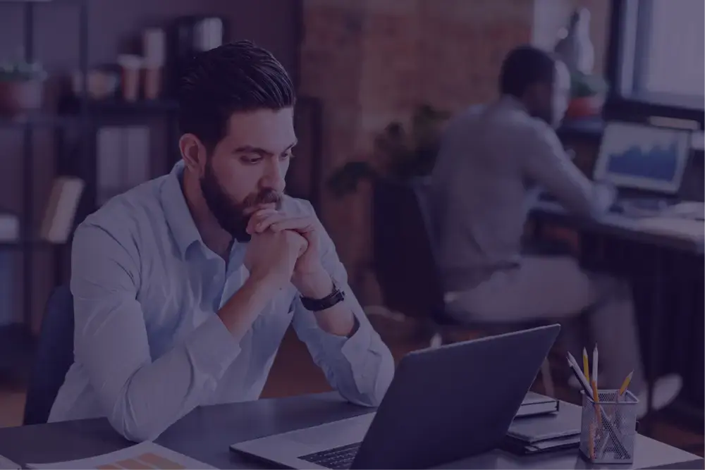 Man with a beard sitting at a desk, looking thoughtfully at a laptop